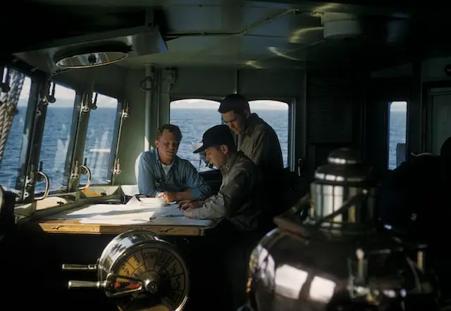Picture of officers discussing something over a navigation chart on a ships bridge 